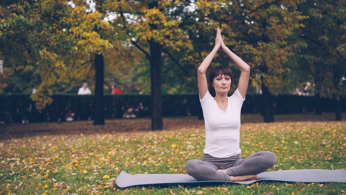 Beautiful girl is concentrated on yoga practice sitting on mat in park in lotus position and bending sideways raising arm. Healthy lifestyle, sports and recreation concept.